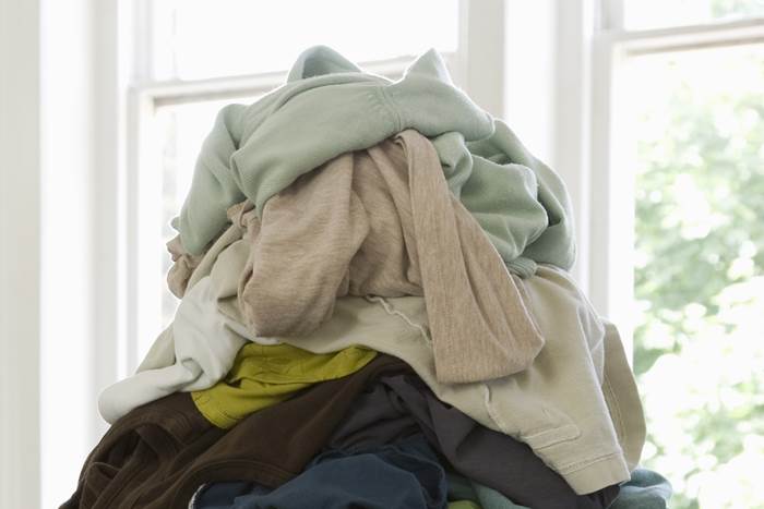 A messy pile of folded and unfolded laundry in muted greens, tans, and blues near a bright window, casual homey mood